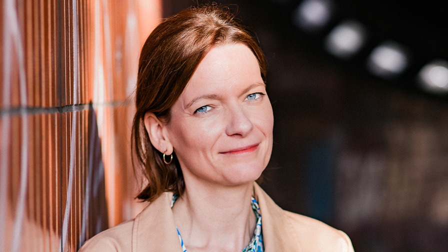 Frau, helle Bluse, braune Haare, an Mauer gelehnt