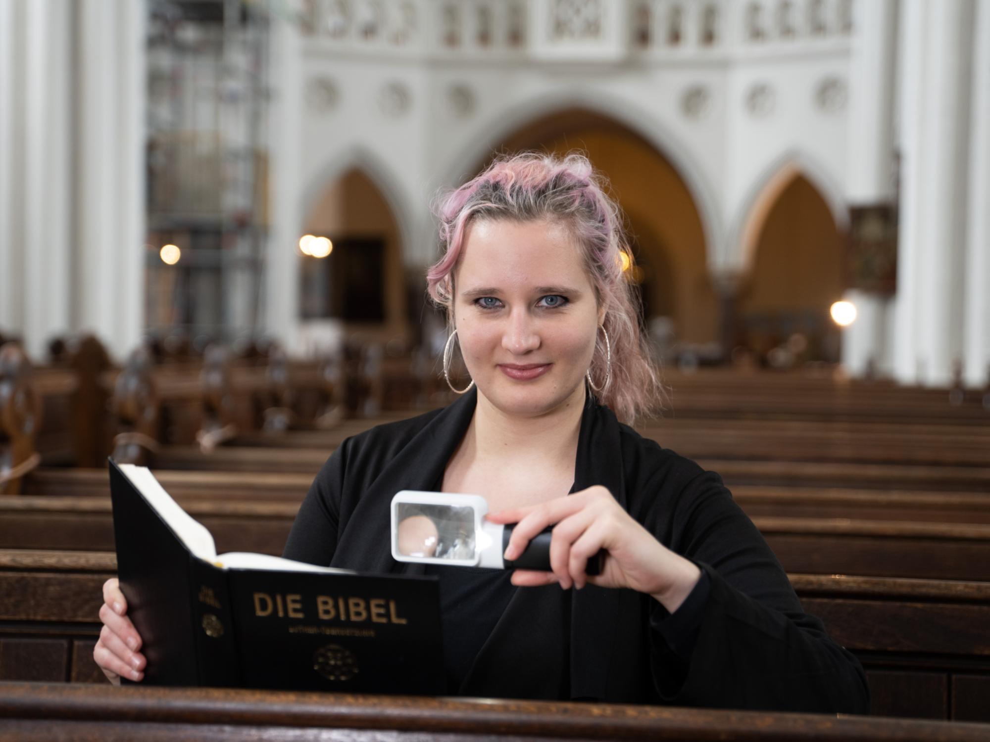 Foto: Eine Frau Mitte 20 sitzt in einer Kirchenbank und lächelt in die Kamera, in einer Hand eine Leuchtlupe, in der anderen eine Bibel. Sie hat langes, dunkelblondes Haar mit rötlichen Strähnchen und trägt große Ohrringe.