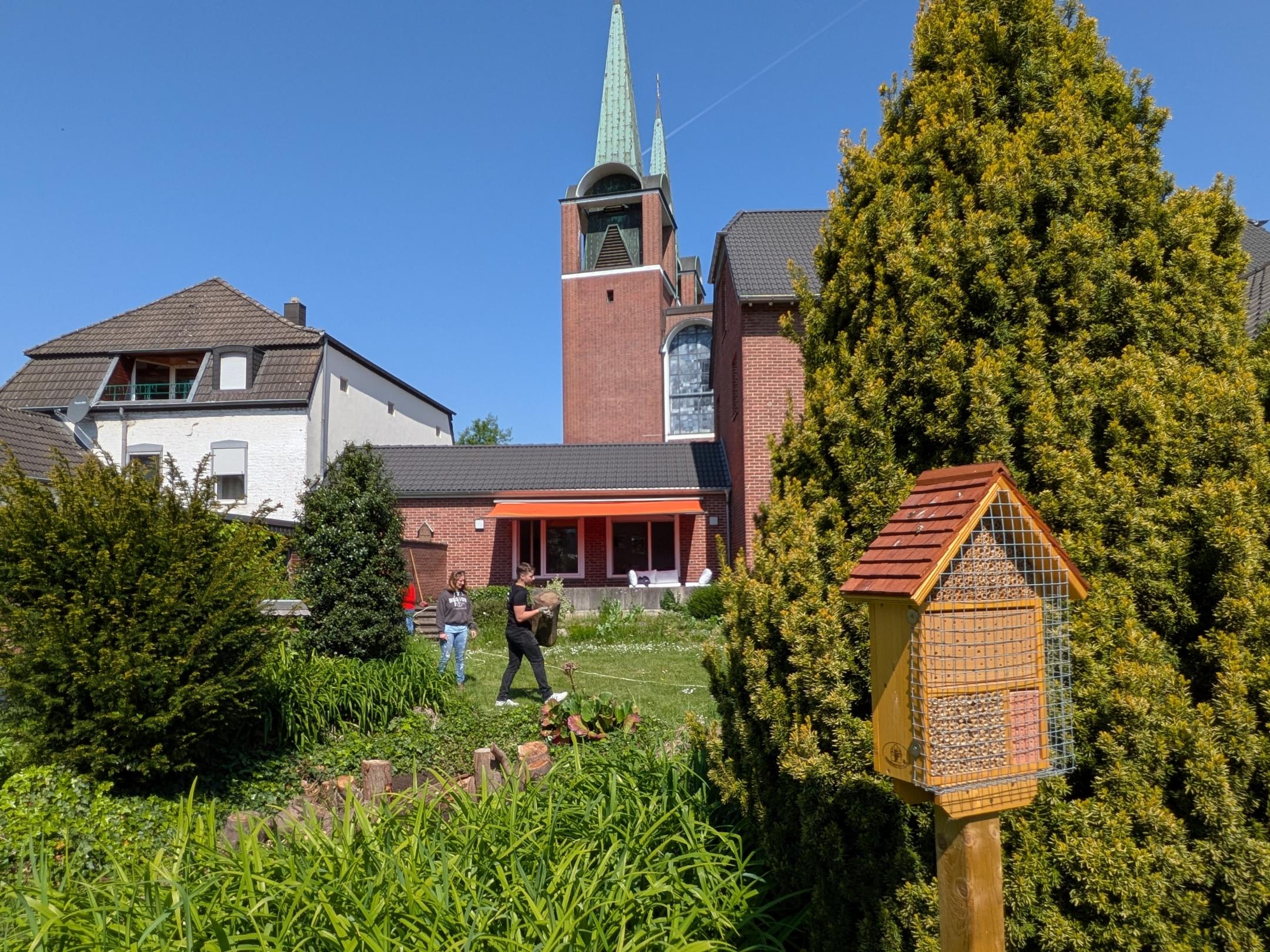 Das Bild zeigt einen Garten mit viel Wiese und einem Vogelhaus. Im Hintergrund sind ein Haus und eine Kirche