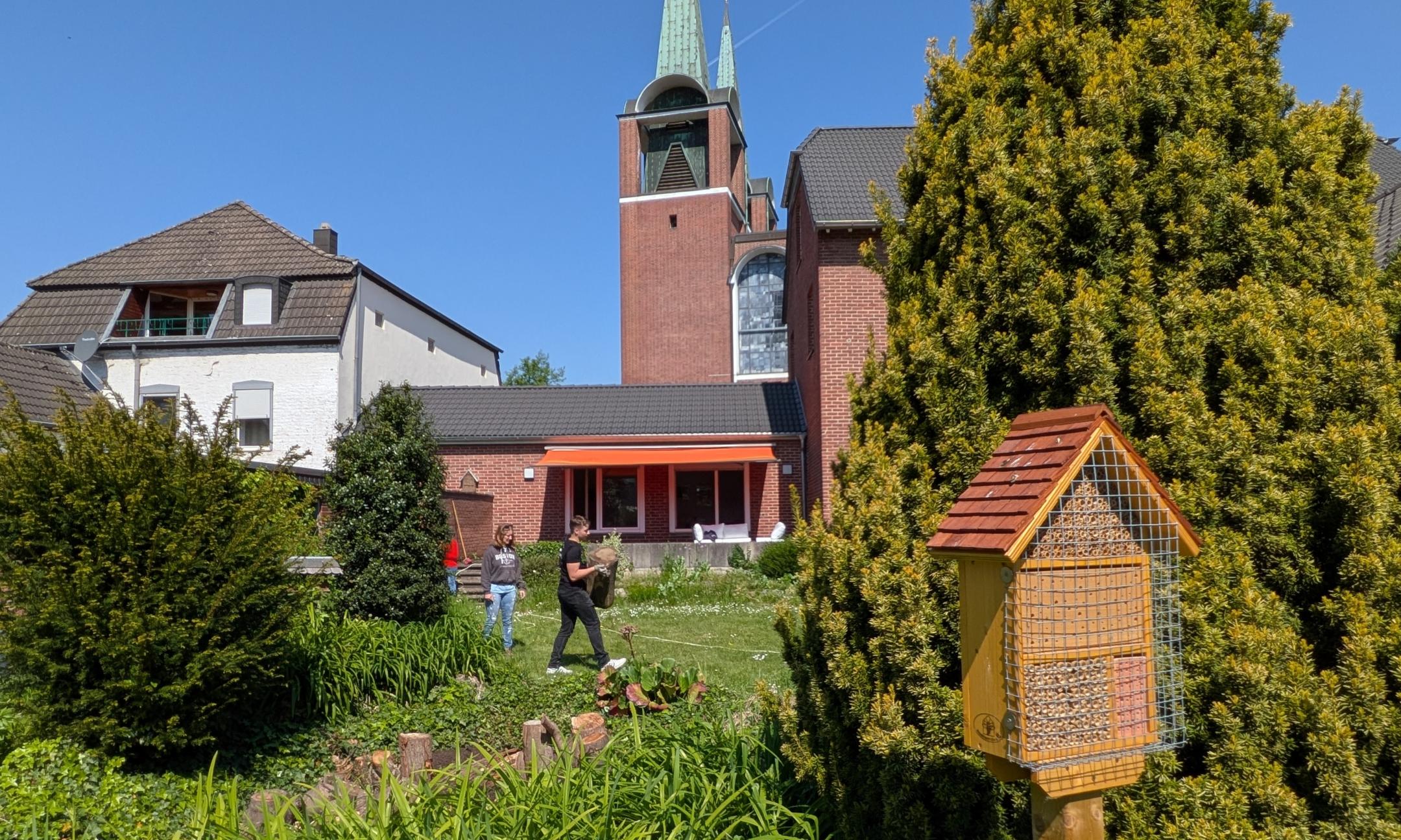 Das Bild zeigt einen Garten mit viel Wiese und einem Vogelhaus. Im Hintergrund sind ein Haus und eine Kirche