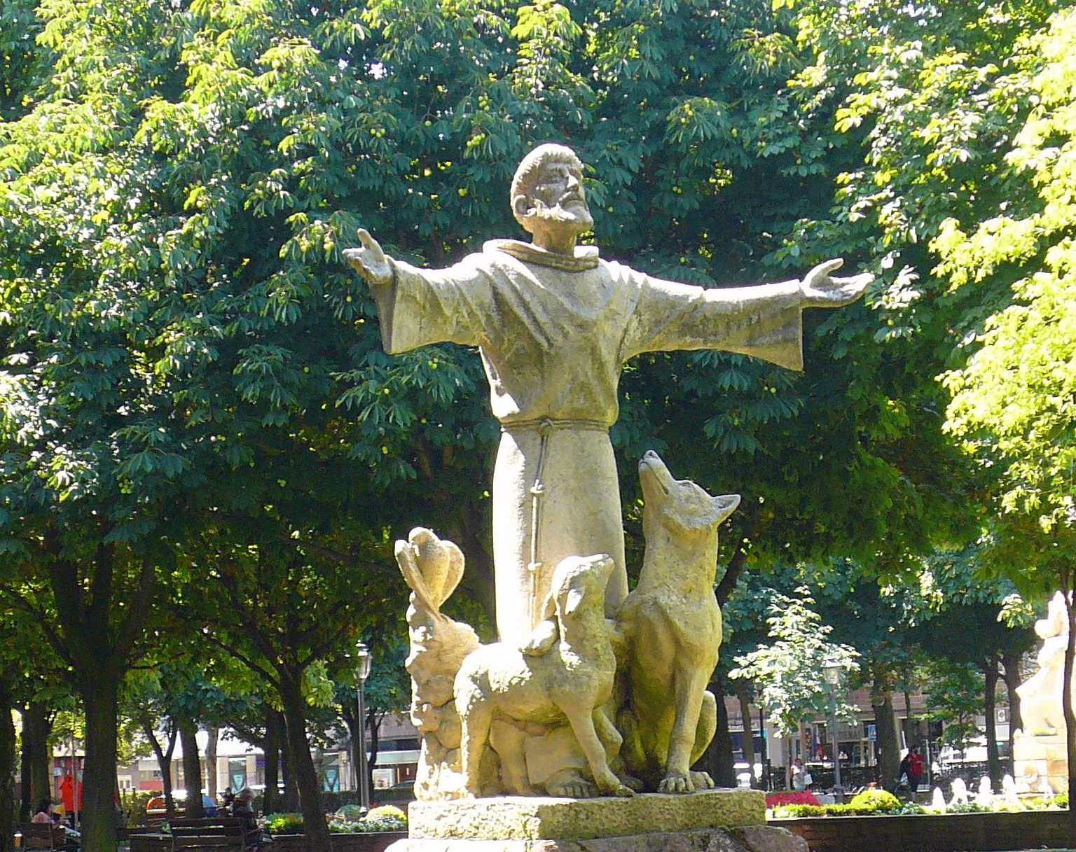 Skulptur des heiligen Franziskus in einem Park vor Bäumen, neben ihm stehen ein Wolf und ein Lamm. Der Heilige hat die Arme weit ausgebreitet.