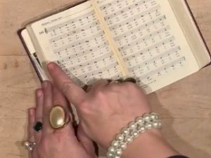 offenes Gebetbuch, eine Frauenhand, die mit Ringen und einem Perlen-Armbband reich geschmückt ist.