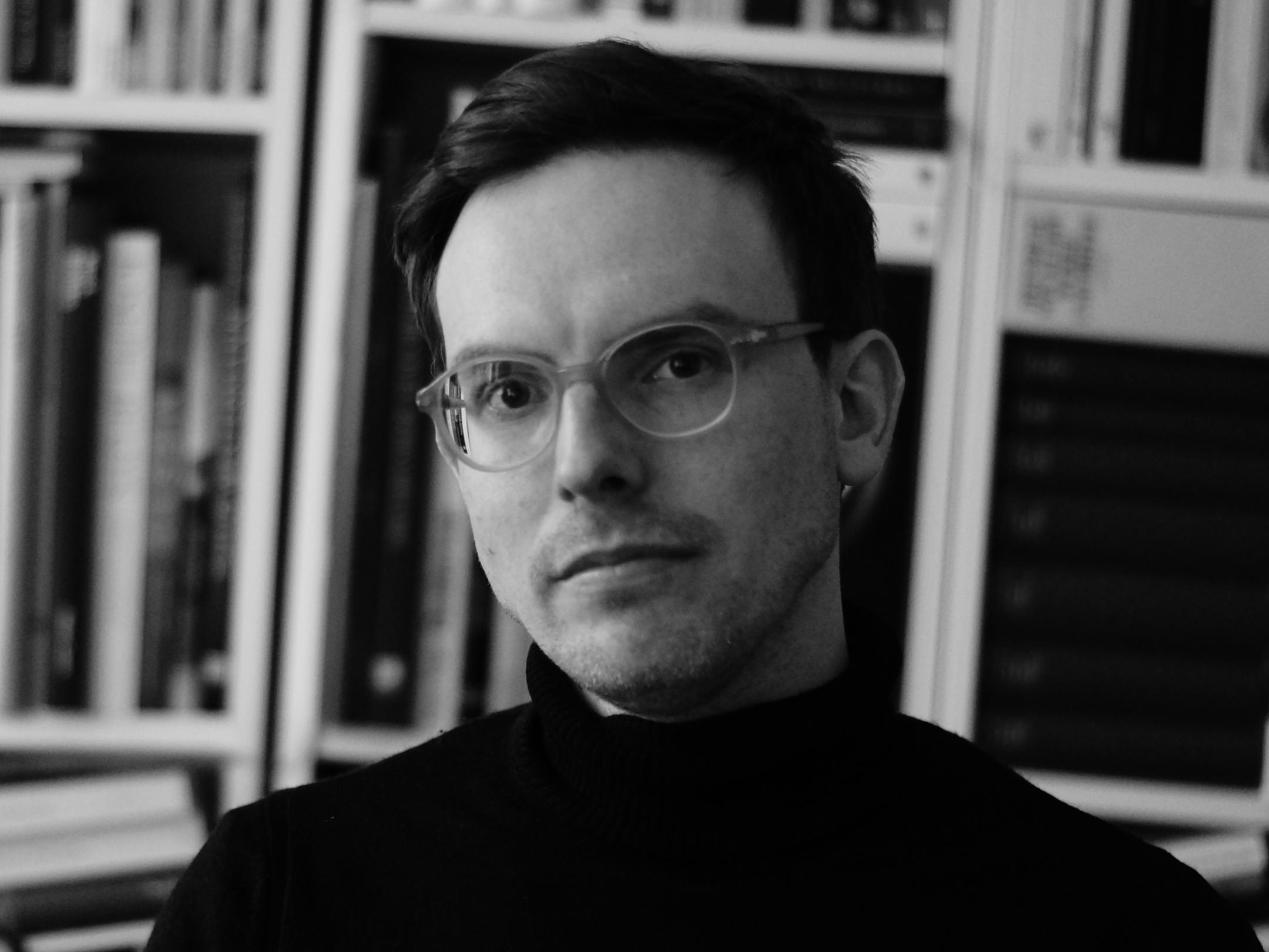 schwarz-weißes Profilbild eines Mannes mit Brille und Kurzhaarfrisur, Mitte 40, vor einem Bücherregal