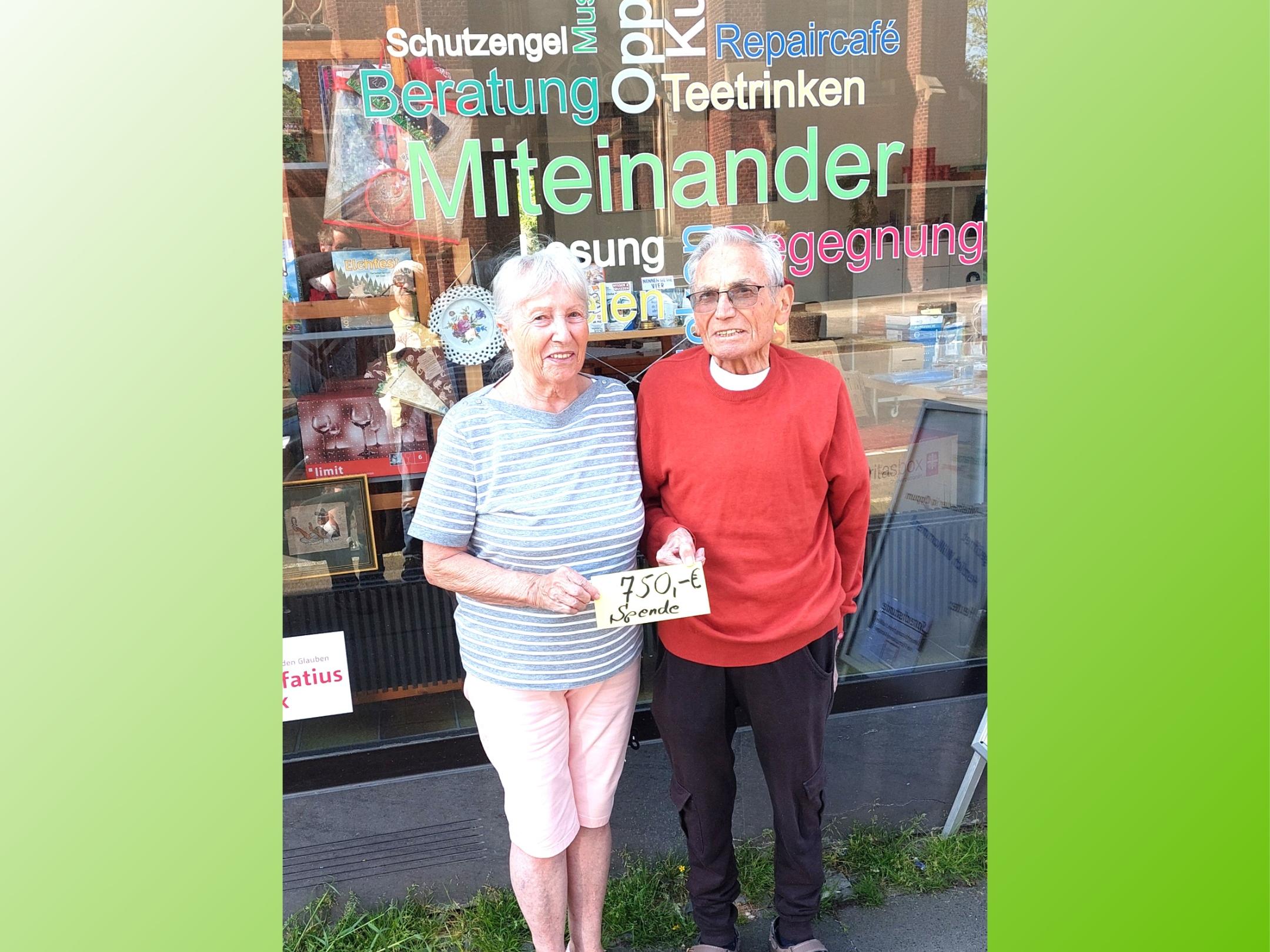 ältere Frau und älterer Mann(= Geschwister) stehen vor dem Schaufenster von MIO in Oppum und halten einen Zettel in der Hand mit 750,- € Spende.