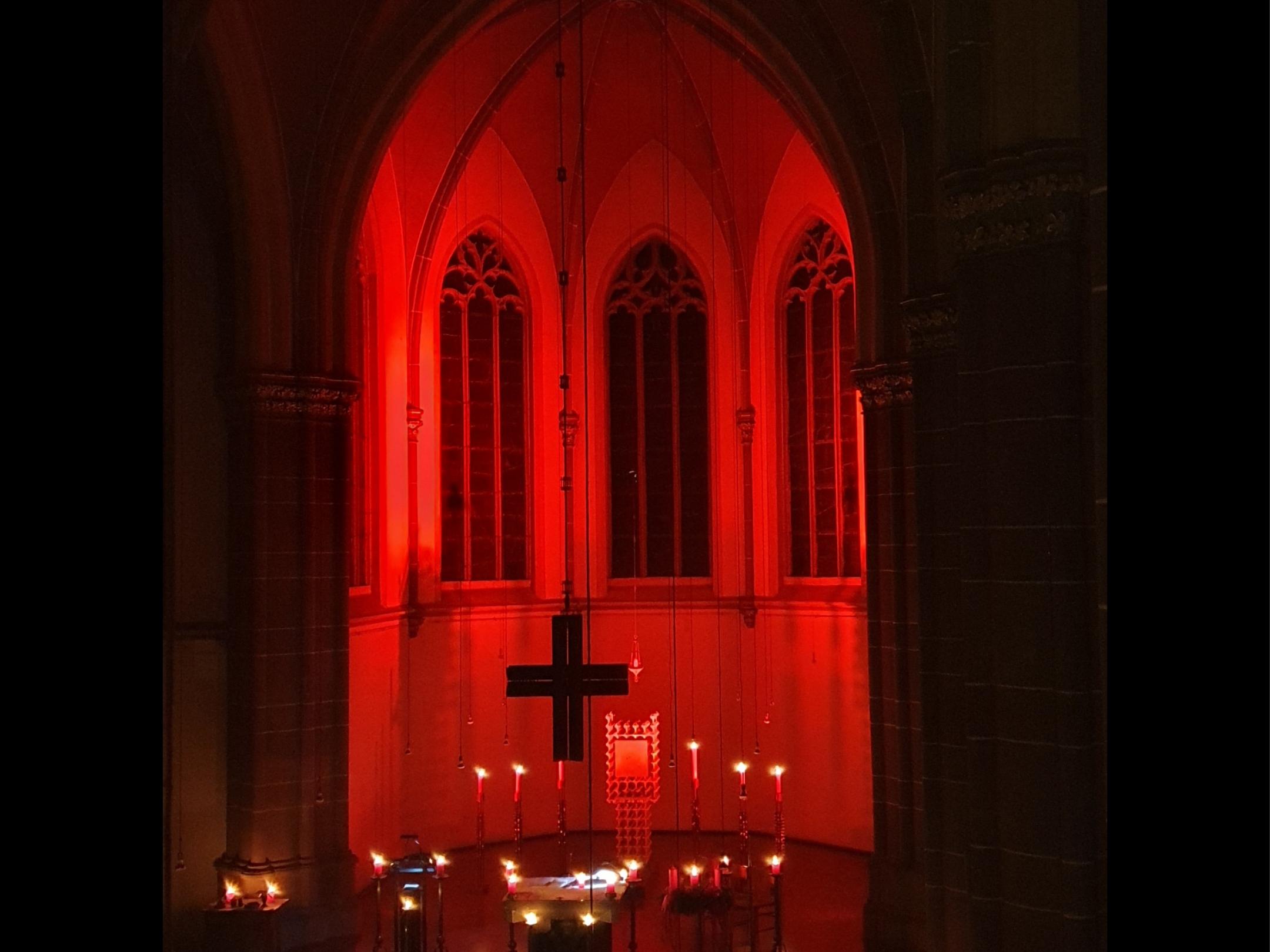 Kirchraum liegt in Dunkelheit nur durch Kerzen, die am Rand der Bänke und vorne im Altar stehen, erleuchtet. Die Apsis wird durch rote Strahler erleuchtet.
